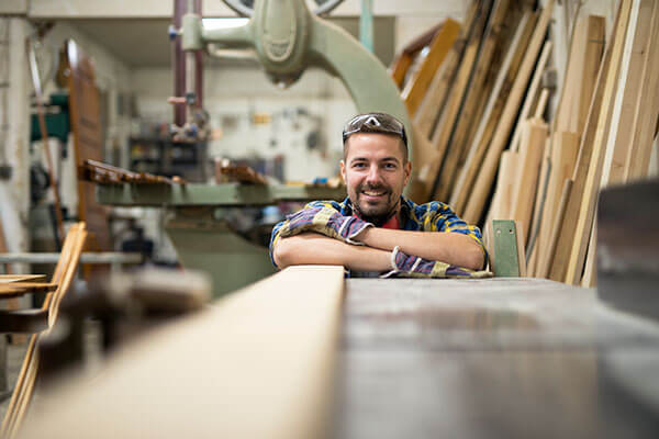 A smiling apprentice in a plaid shirt and safety goggles leans on a workbench in a woodshop, surrounded by lumber stacks and woodworking equipment.