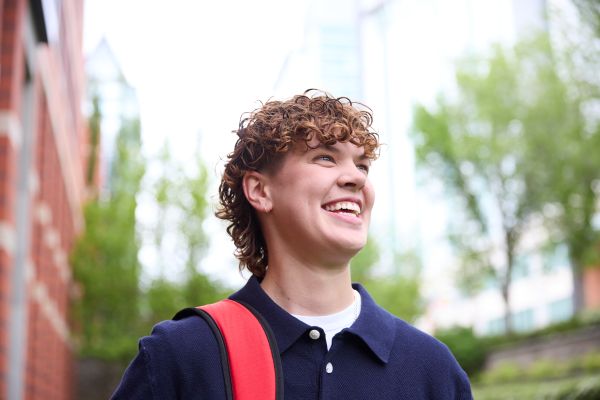 Person with a backpack stands outdoors near modern buildings and trees.